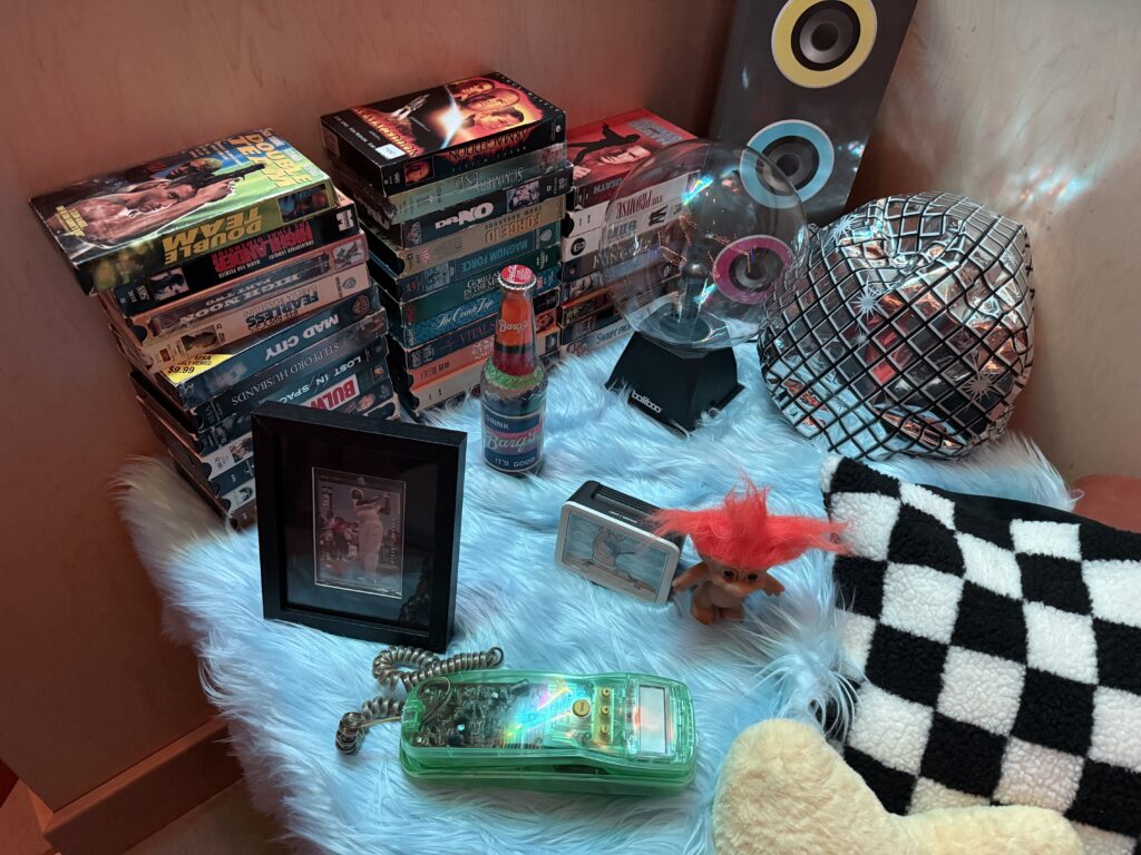 A table with stacks of VHS tapes, a troll doll, and a greeen transparent telephone. 
