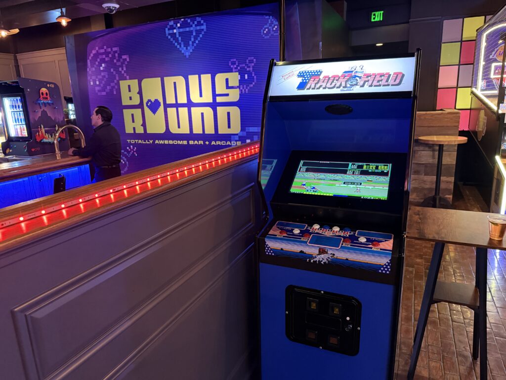 Arcade area with a blue Track & Field game cabinet, a wooden bar counter, and a man standing at the counter under colorful lights.