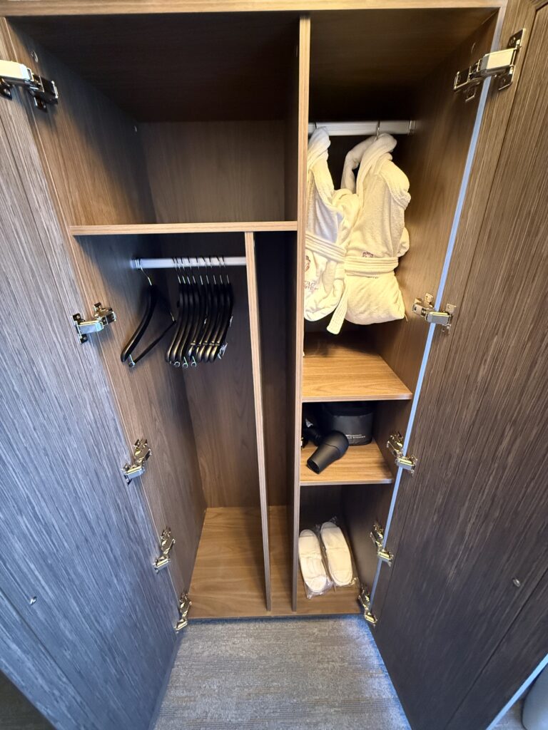 Open wooden wardrobe with white bathrobes hanging on the right and a lower shelf holding slippers and a hairdryer nearby.