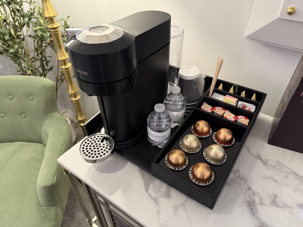 Black espresso machine on a marble counter with a capsule drawer and two water bottles nearby in a lounge area.