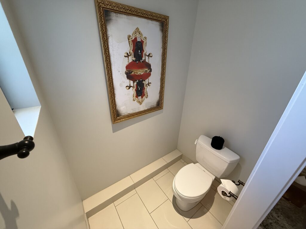 Small bathroom with two white toilets side by side, beige walls, tiled floor, and a large ornate framed picture on the wall.