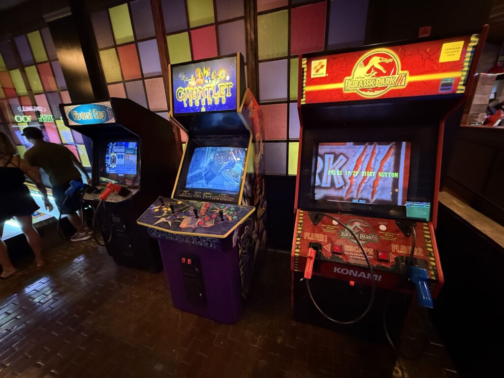 Row of classic arcade game cabinets in a dimly lit arcade, featuring Gauntlet Legends and Jurassic Park, with colorful tiled wall behind.