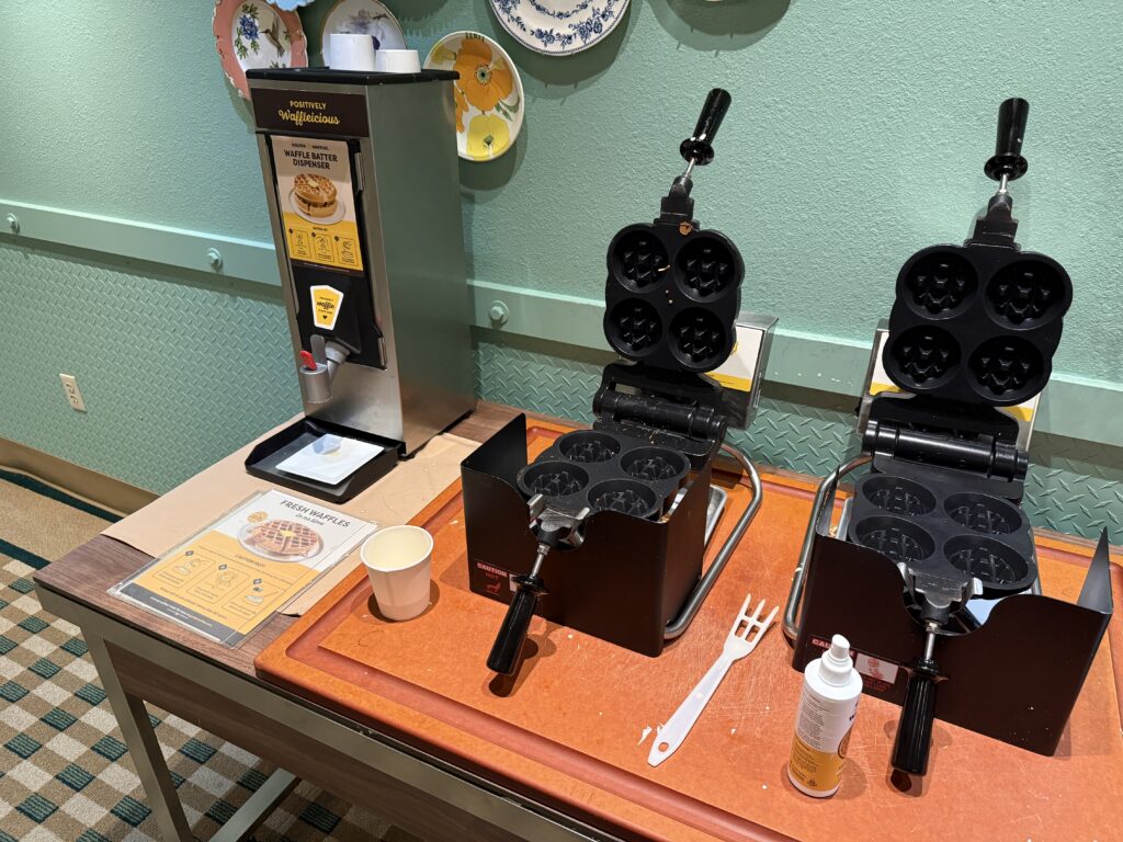 A waffle batter dispenser next to two waffle irons. 