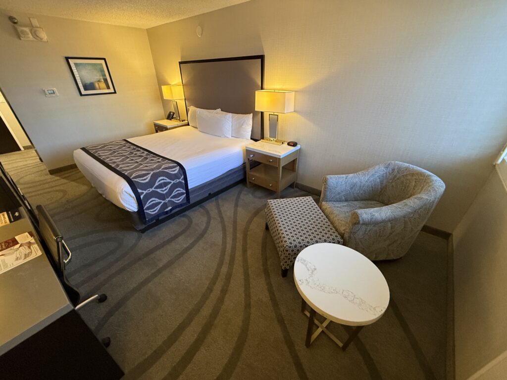 A small chair and circular end table in the foreground, with a king bed flanked by lamps in the background. 