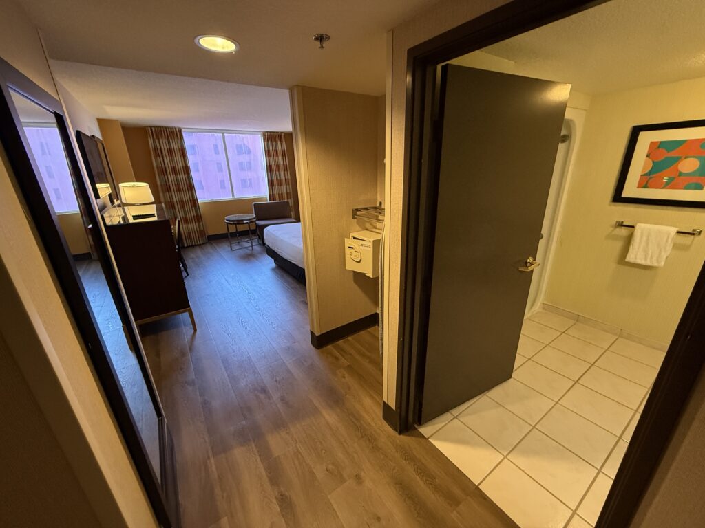 Hotel room interior with a bed, seating area by a window, and a bathroom doorway visible from the hallway viewed at an angle.