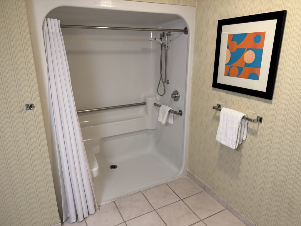 Shower/tub combo with a metal grab bar, handheld shower and white curtain. A towel rack stands ready to the right.