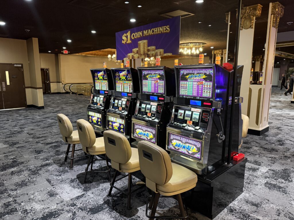 Row of electronic slot machines with beige chairs in a casino lounge, under a large purple banner that reads '$1 COIN MACHINES'.
