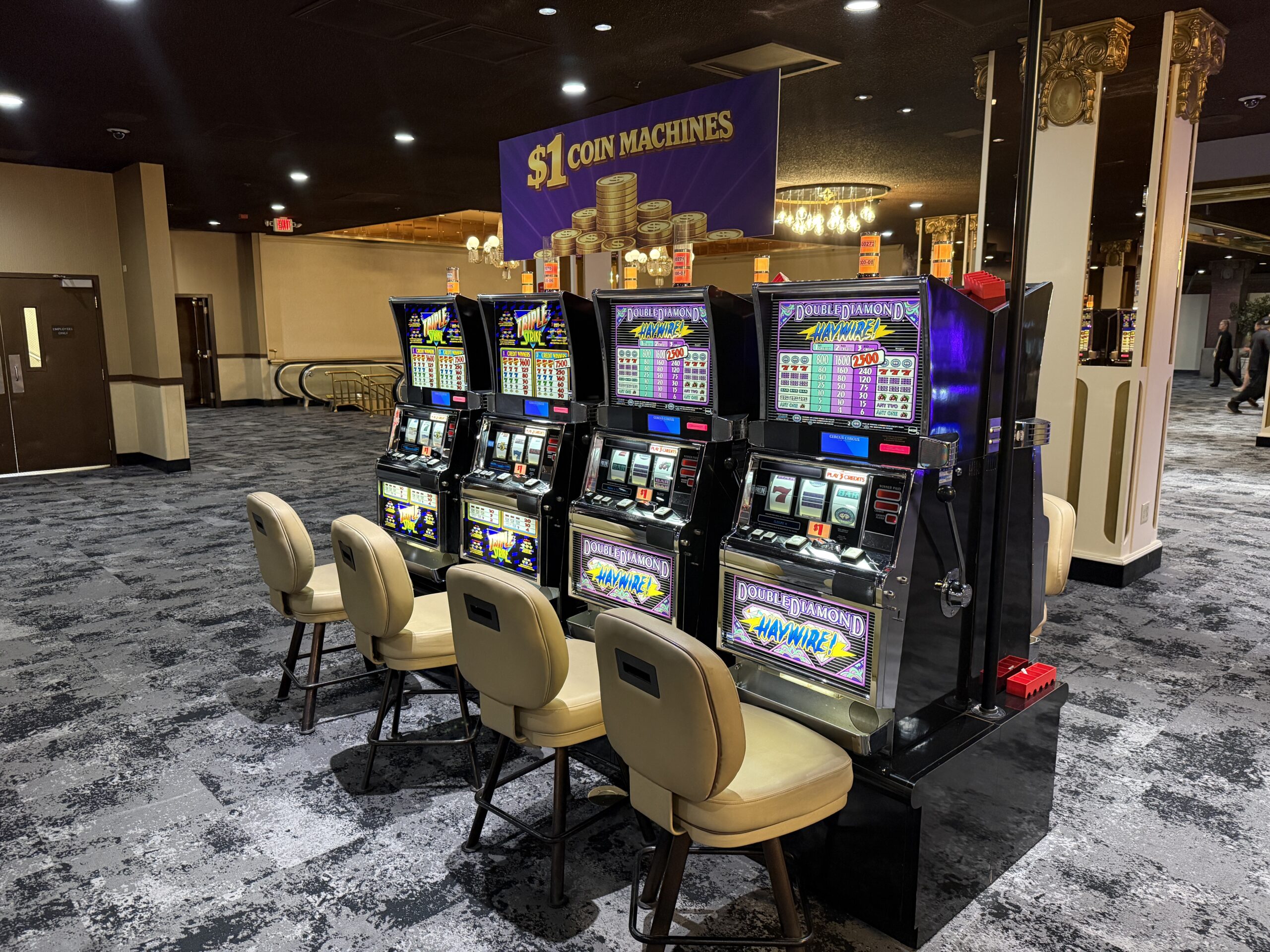 Row of electronic slot machines with beige chairs in a casino lounge, under a large purple banner that reads '$1 COIN MACHINES'.