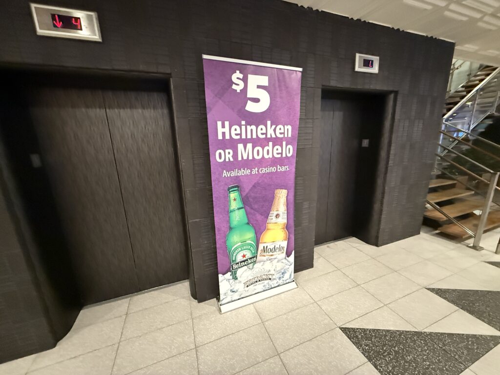 Purple promotional banner reading '$5 Heineken or Modelo' with beer bottles at the bottom, between two elevator doors in a dark hallway area.