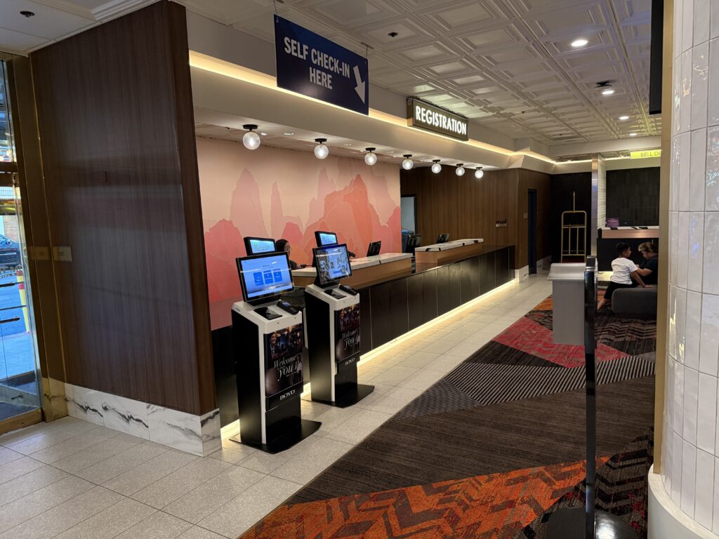 Hotel lobby registration desk with self check-in sign and computer kiosks and a standard employee manned desk. 