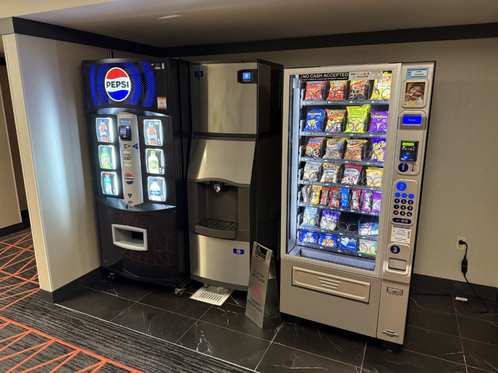 Two vending machines selling drinks and snacks on either side of an ice dispenser. 