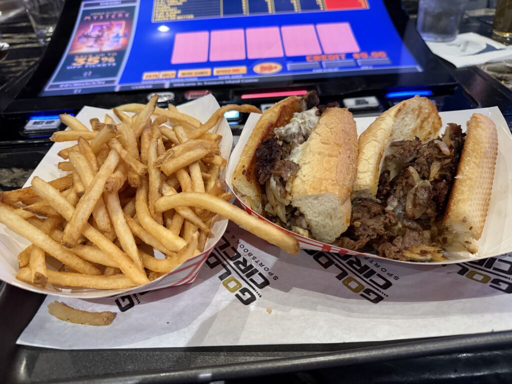 A philly cheesesteak and french fries on a counter in front of a bartop video poker machine.