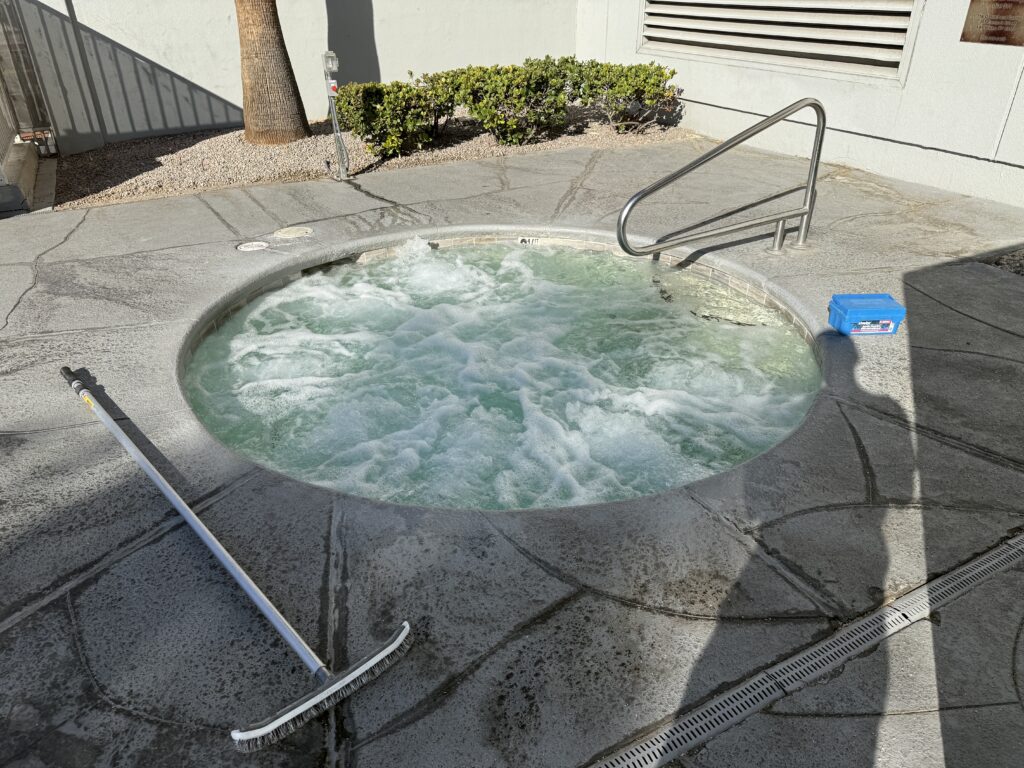 Circular hot tub with bubbling water on a concrete deck; a pool brush and pole lie nearby on the ground.