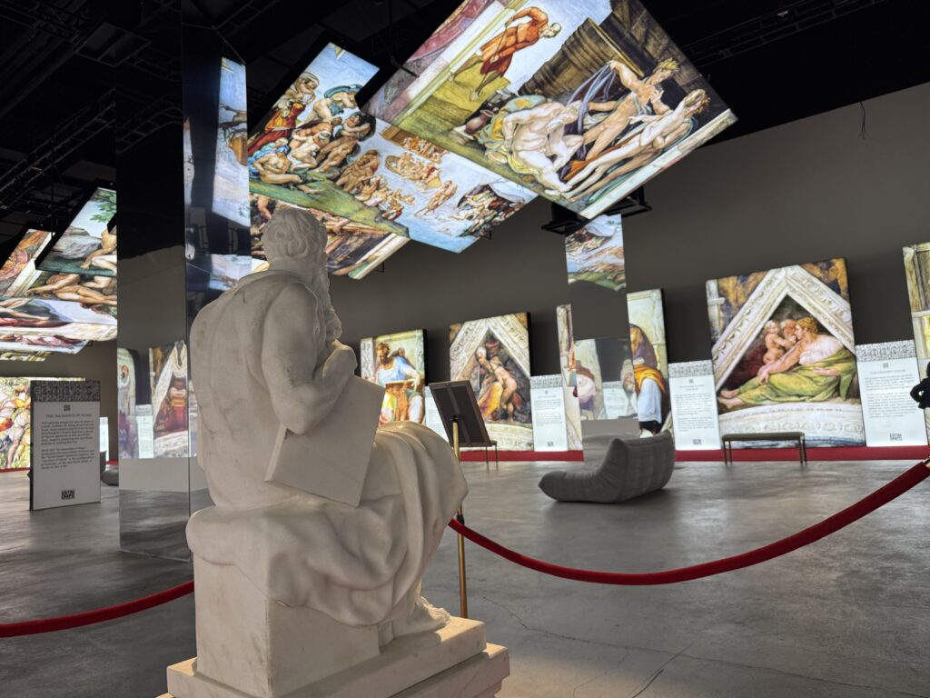 A white stone statue in the foreground, with illuminated paintings in the background. 