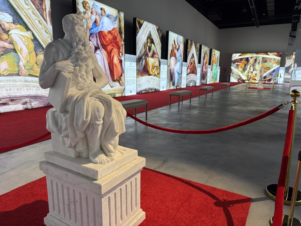 A white statue on a red carpet. In the background are illuminated frescoes. 