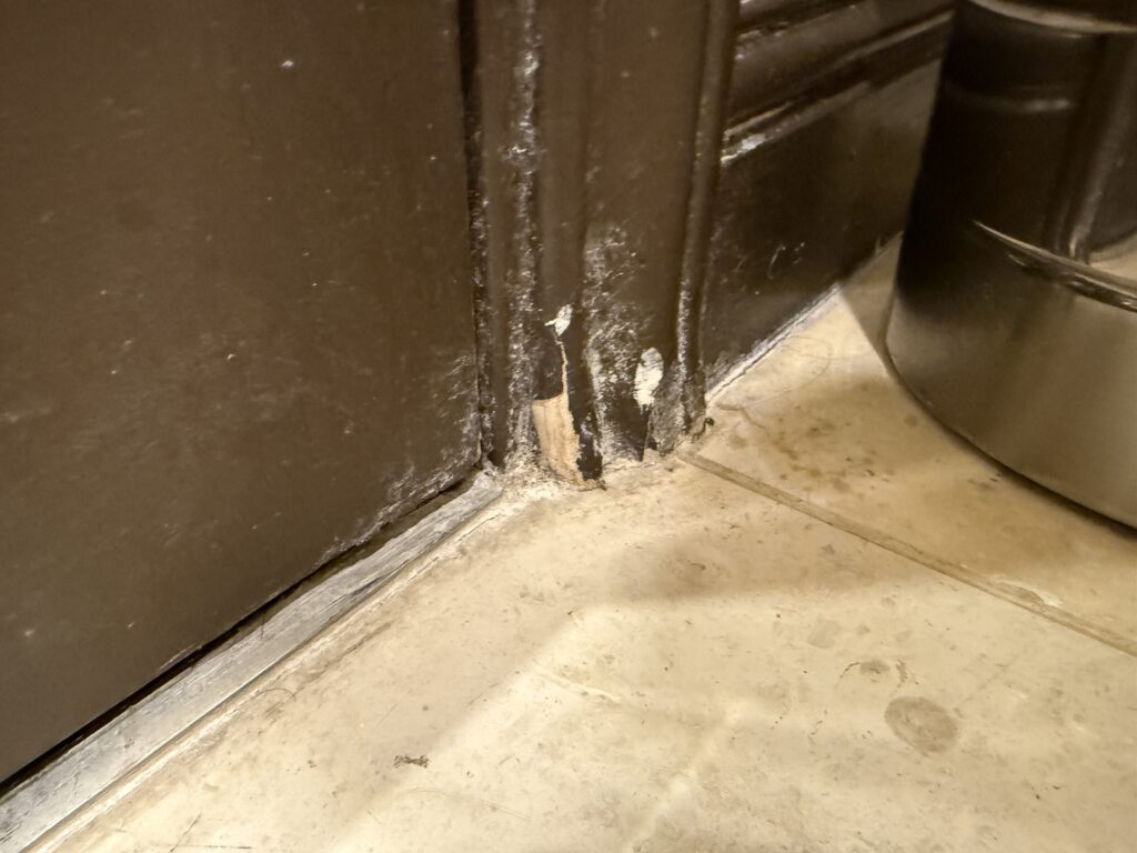 Close-up of a brown door frame with chipped paint and a dusty floor nearby.