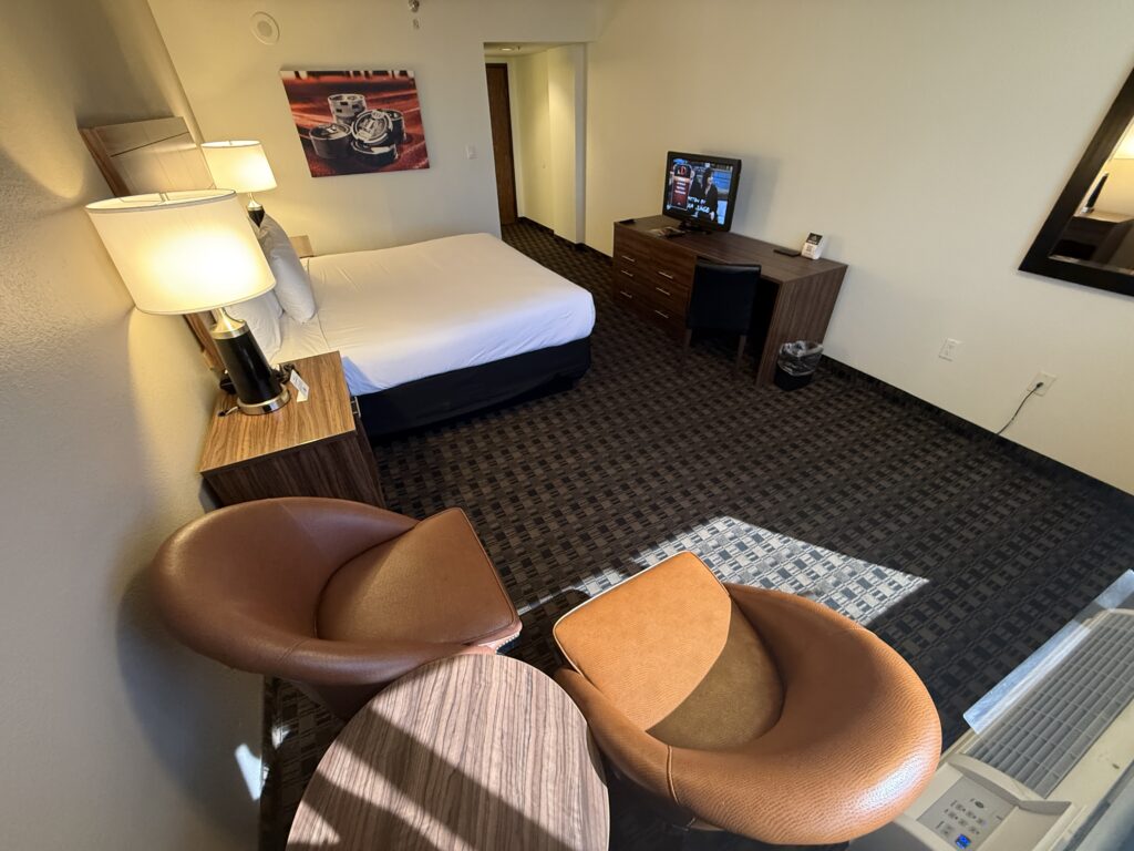 A circular table and two brown faux leather chairs in the foreground. In the background the king bed is pointed at the tiny TV.