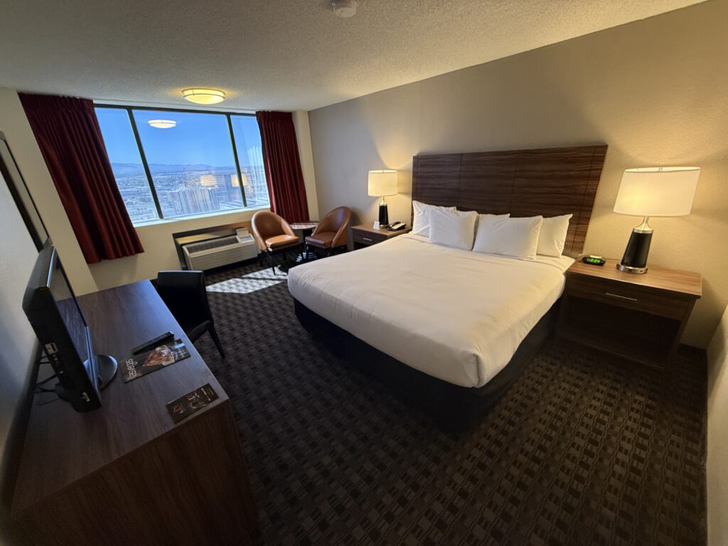 A wide angle that shows a king bed, basic faux wood headboard, and window in the background.
