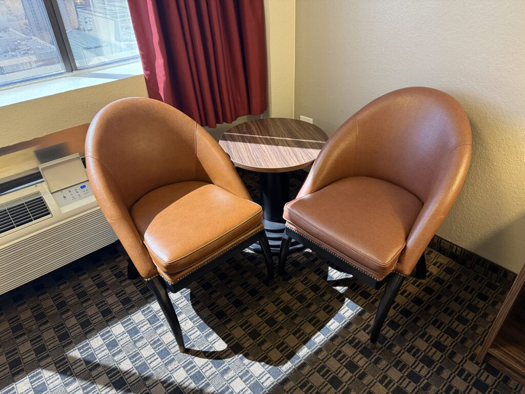 Two brown leather chairs surround a small round wood table by a window with red curtains.