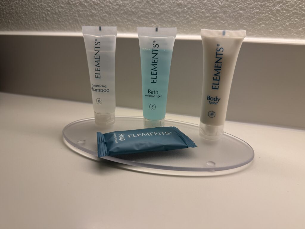 Three ELEMENTS toiletries—left: shampoo, center: bath gel, right: body lotion—arranged on a bathroom counter with a blue sachet in front.