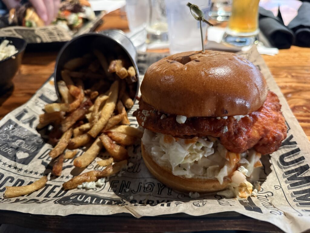 A buffalo chicken sandwich next to a cup full of french fries. 