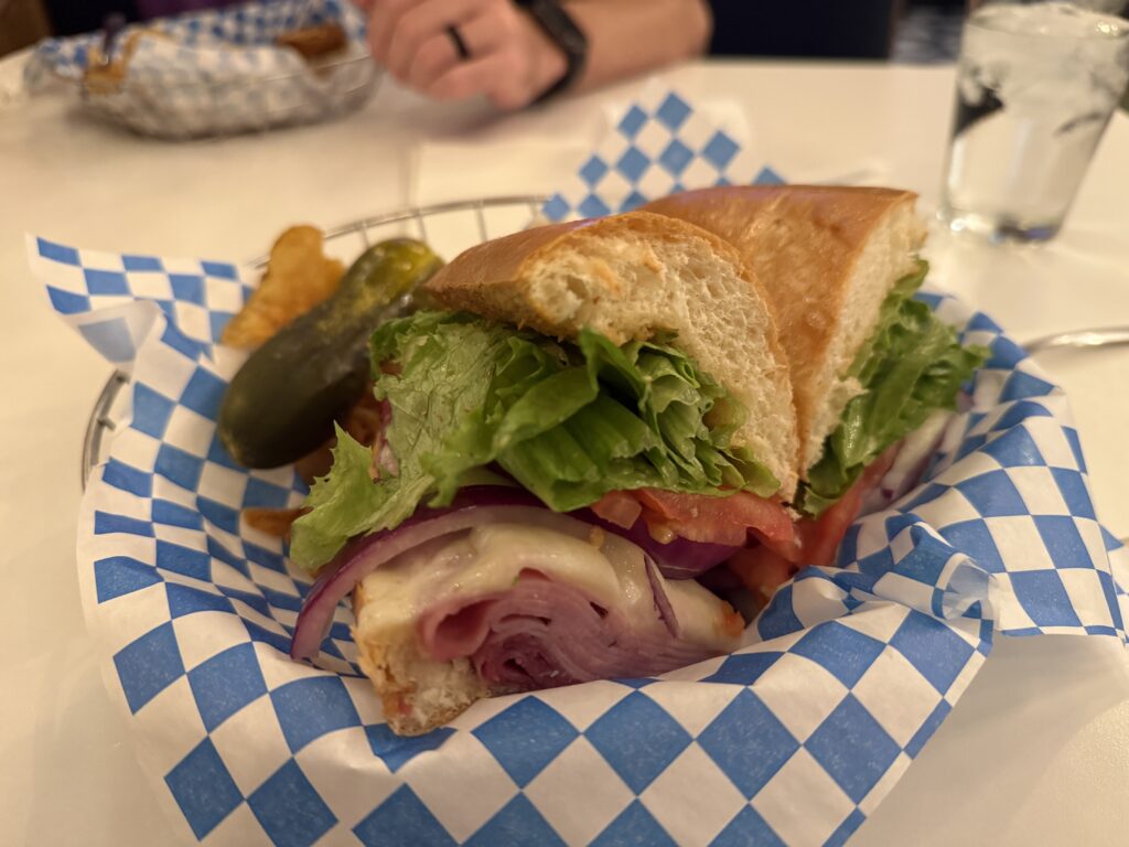 Close-up of a sandwich with lettuce, tomato, onions, and sliced meat on a soft bun in a blue-checkered paper liner in a basket with a side of pickles and a glass nearby (informative).