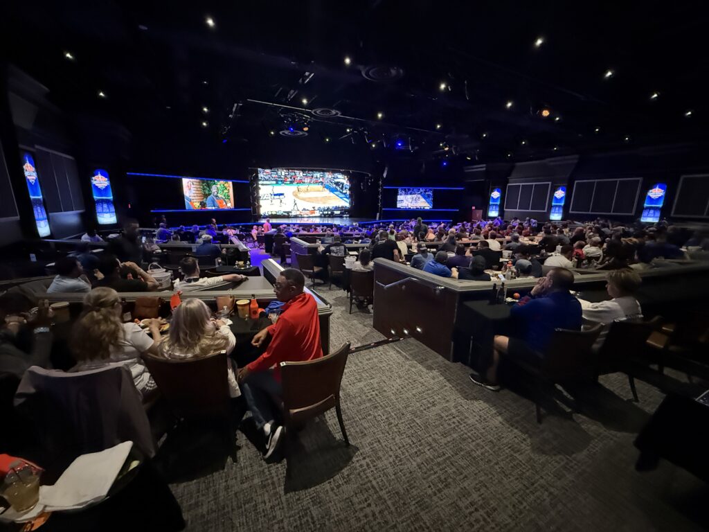 A full theater of people, all looking at giant screens playing college hoops.