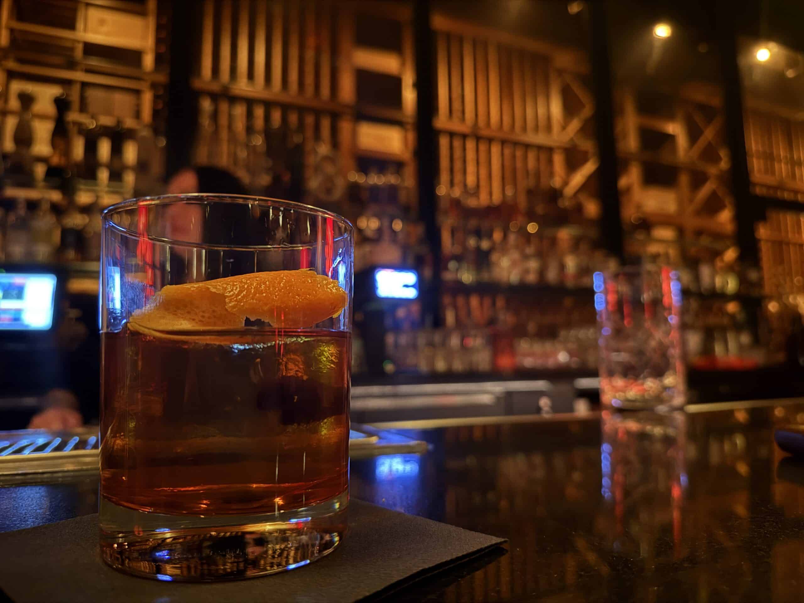 An old fashioned, in focus in the foreground. The background is the illuminated wine rack.