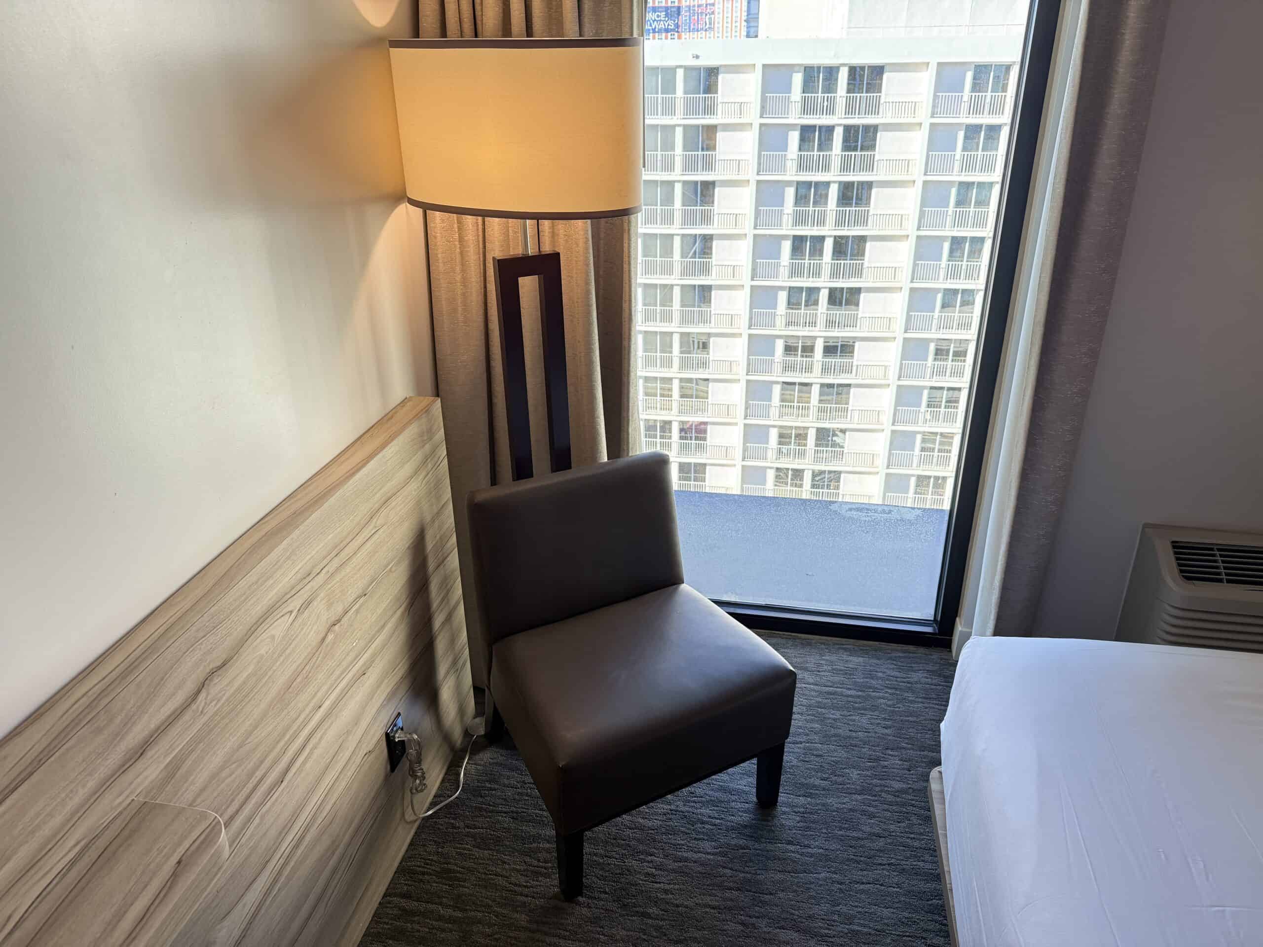 A brown, basic chair and lamp next to a tall, skinny window.