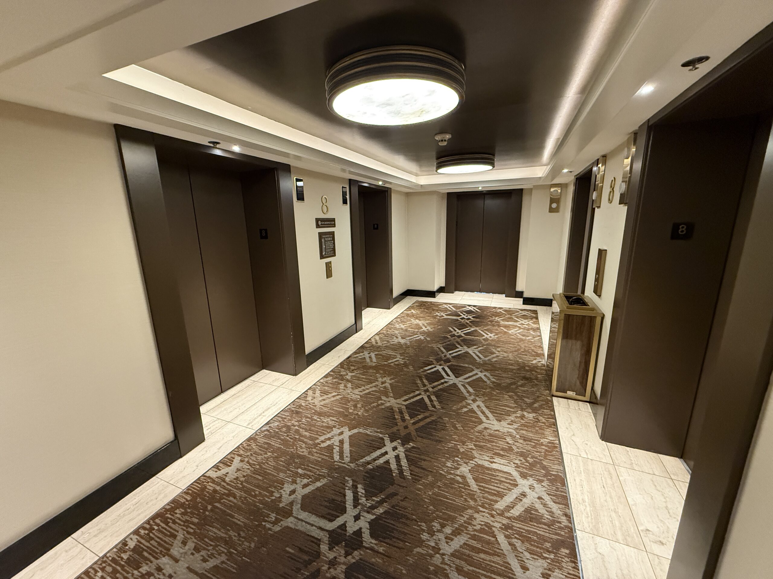 A well lit elevator lobby with creme walls and brown accents.