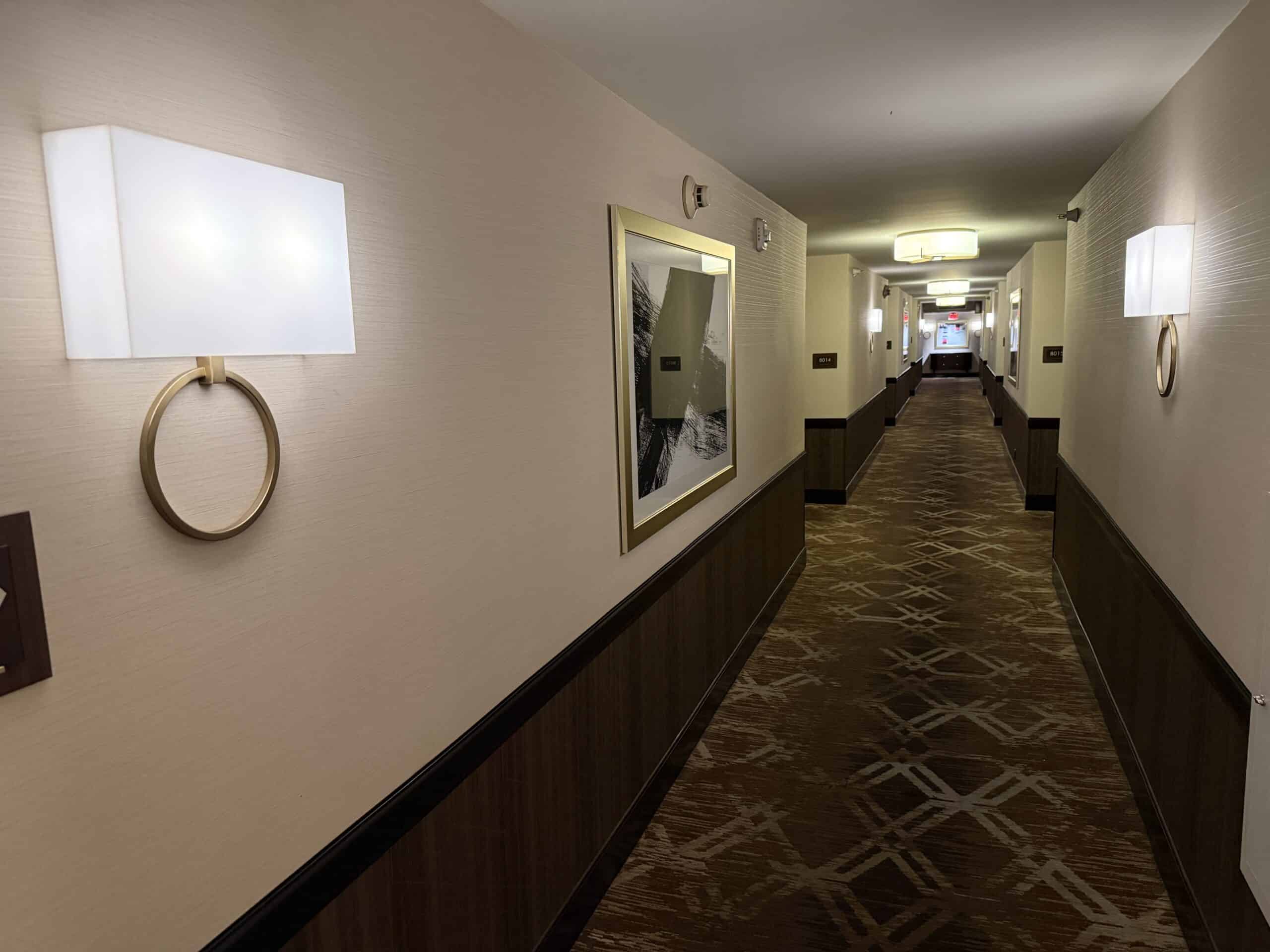 A well lit, fashionable hotel hallway with artwork, creme walls, and brown carpet.