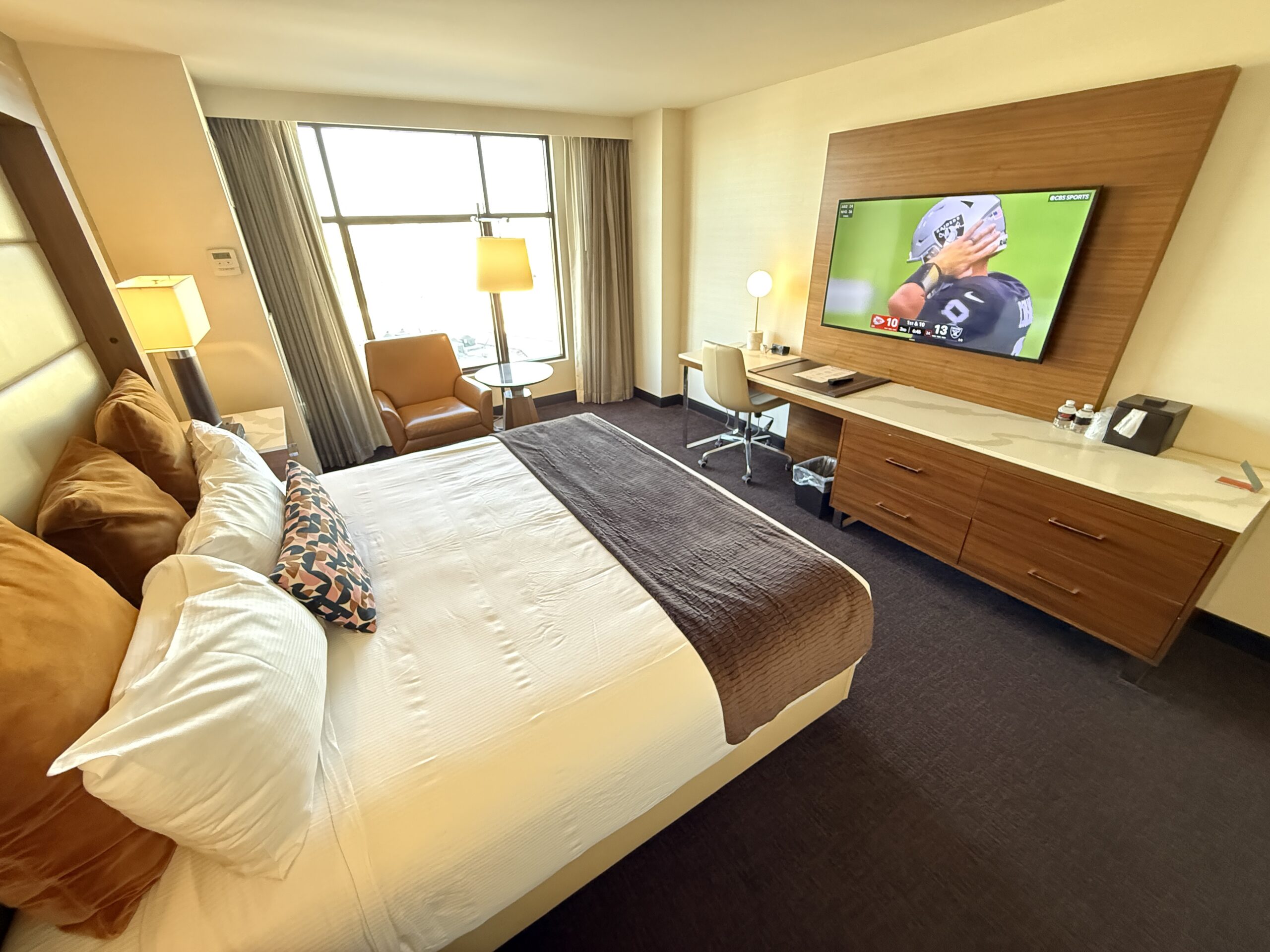 A wide angle view of the room that shows the bed pointed at the TV with the window and a chair in the background.