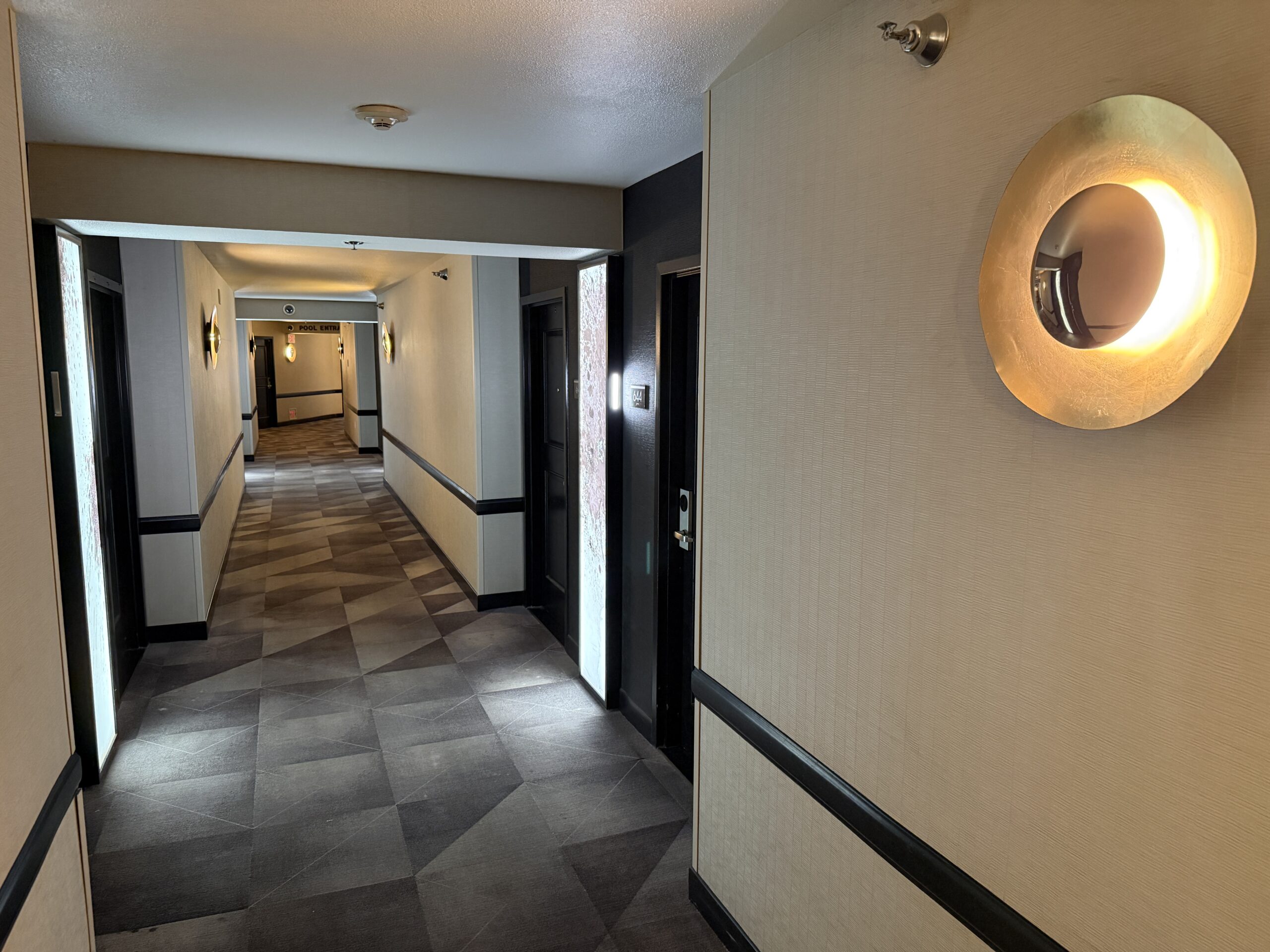 A hotel hallway, with dark carpet, and unique lighting.