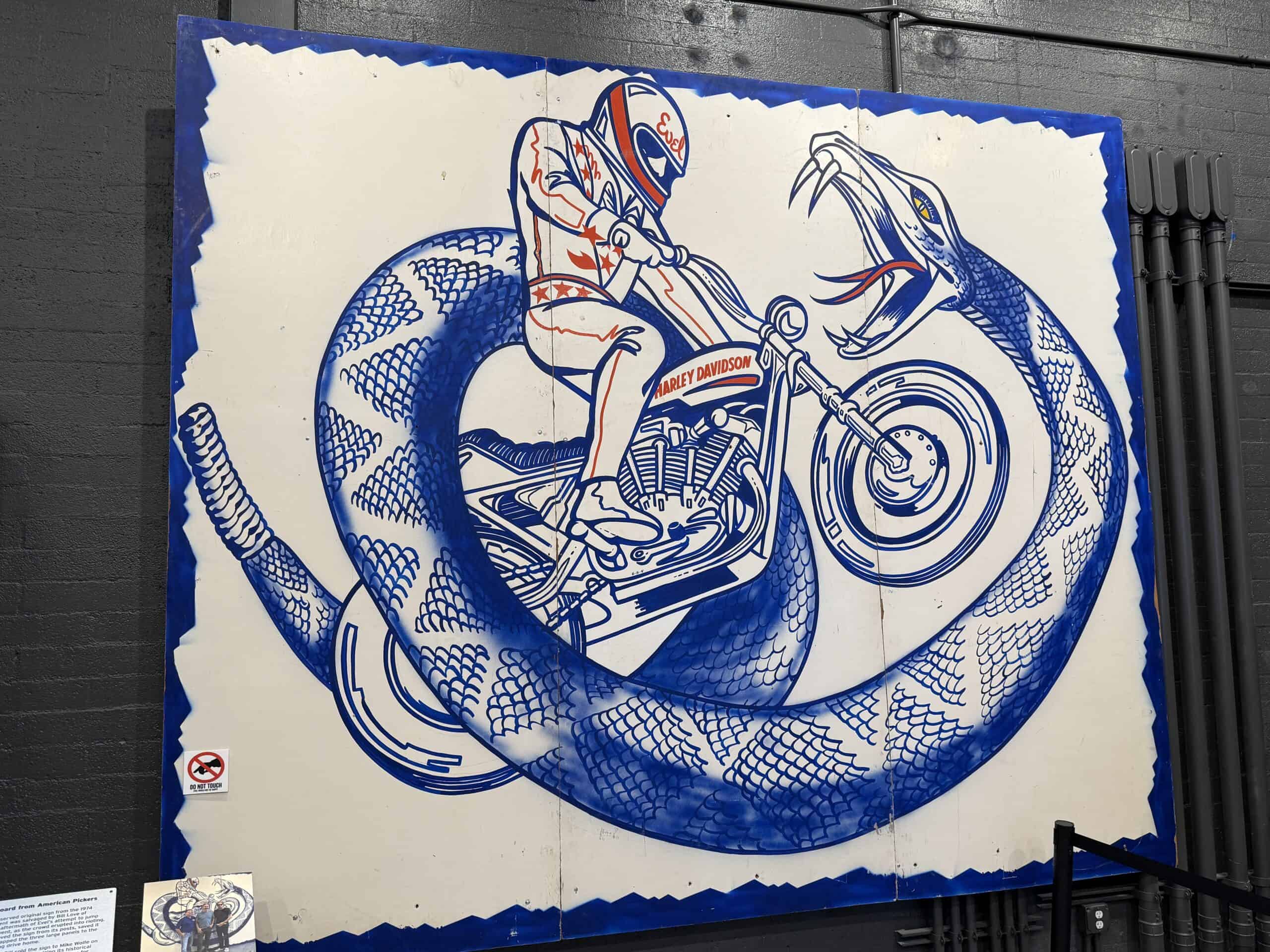 A billboard for the Snake River Canyon jump depicting a blue snake with Evel on a Mororcycle jumping through the snake's body.